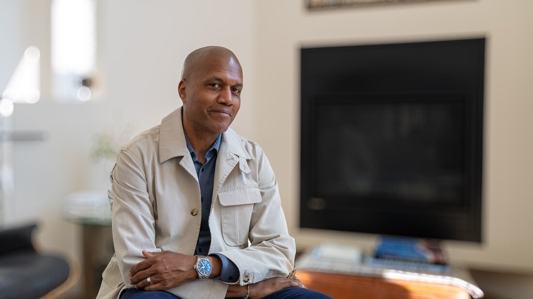 Kevin Bethune in tan jacket sitting in front of fireplace.