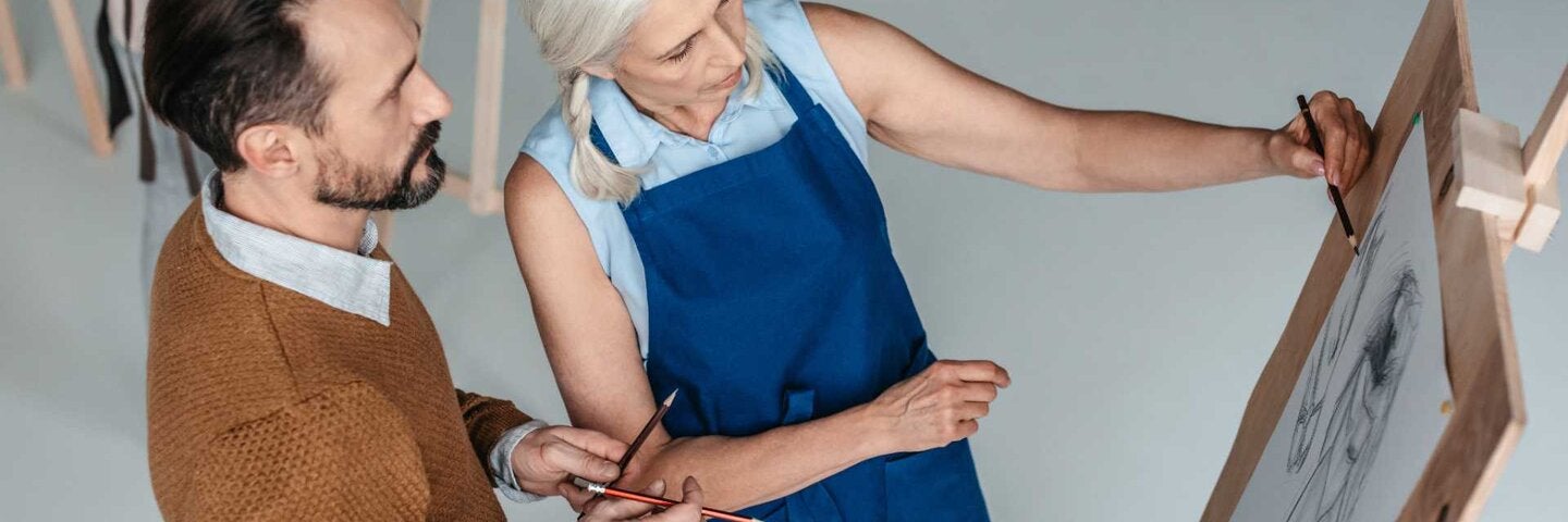 A mature man holding pencils and looking at senior woman drawing on easel