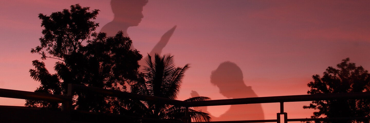 looking out from a deck at sunset. Trees are black silhouettes. One person reading, one writing.
